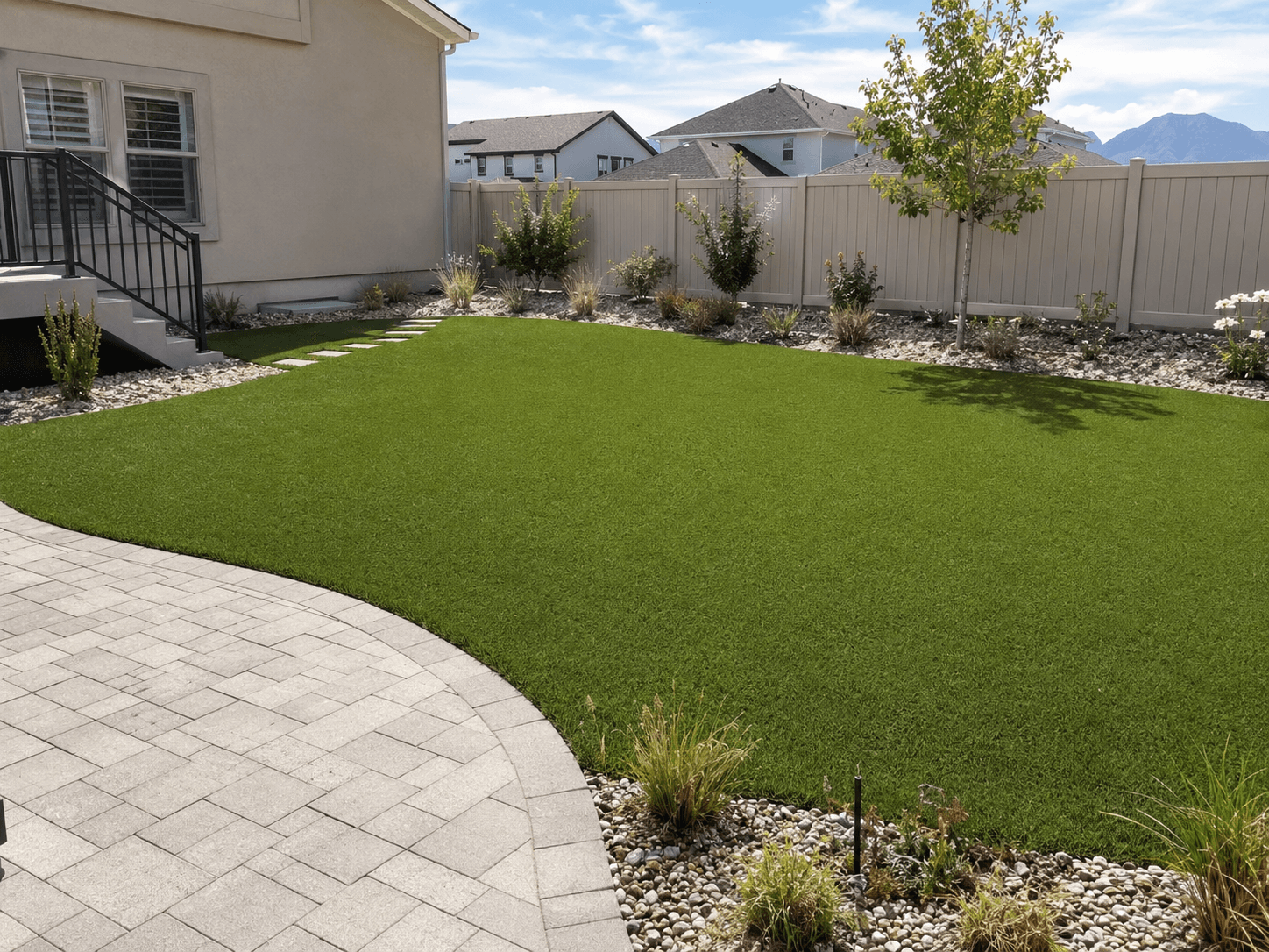 Backyard artificial turf lawn with a clean paver edge, rock border planting beds, and a stepping-stone path along a vinyl privacy fence