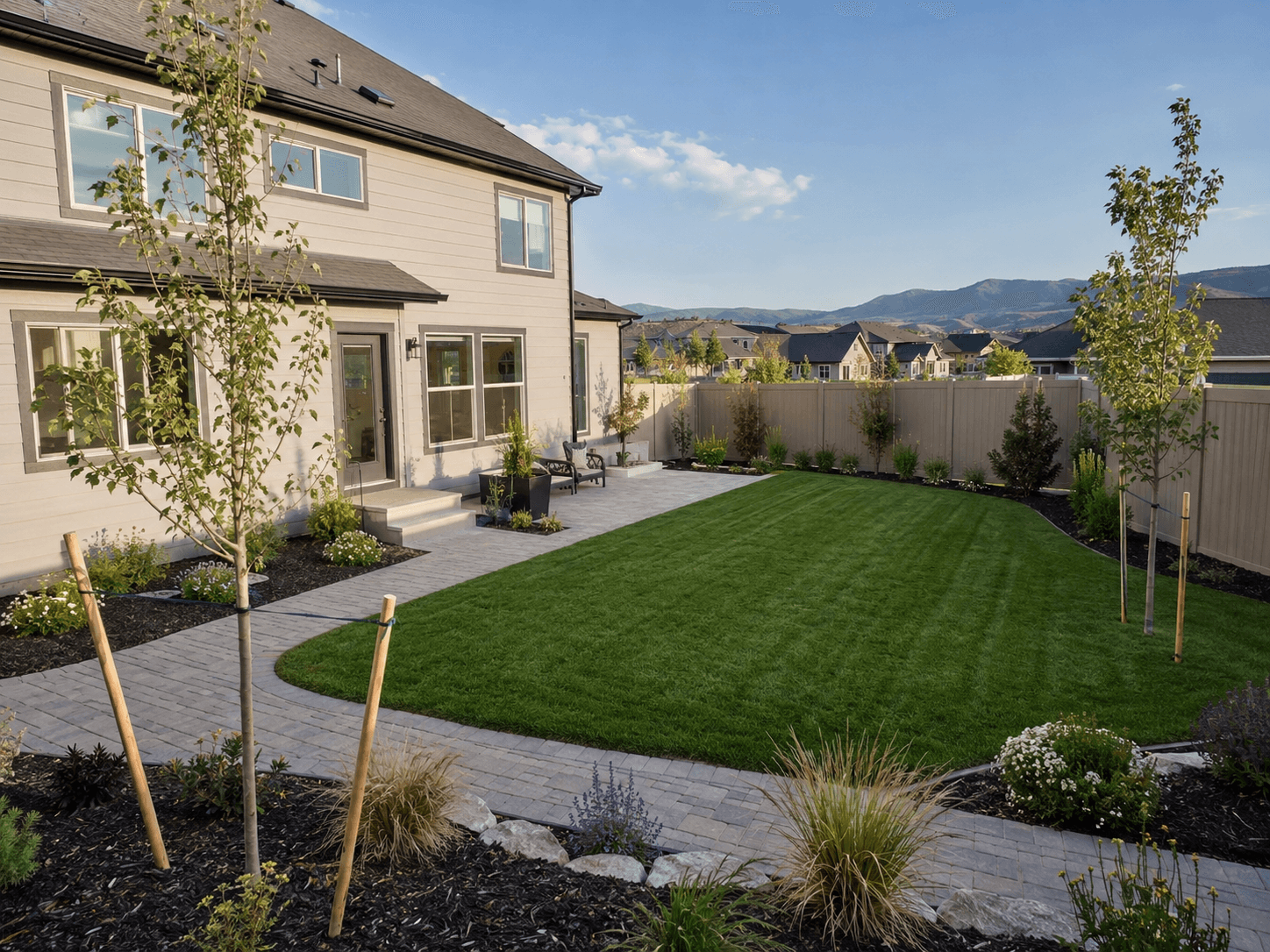New construction backyard with a fresh sod lawn, staked young trees, a paver patio, and a vinyl privacy fence, mountains behind