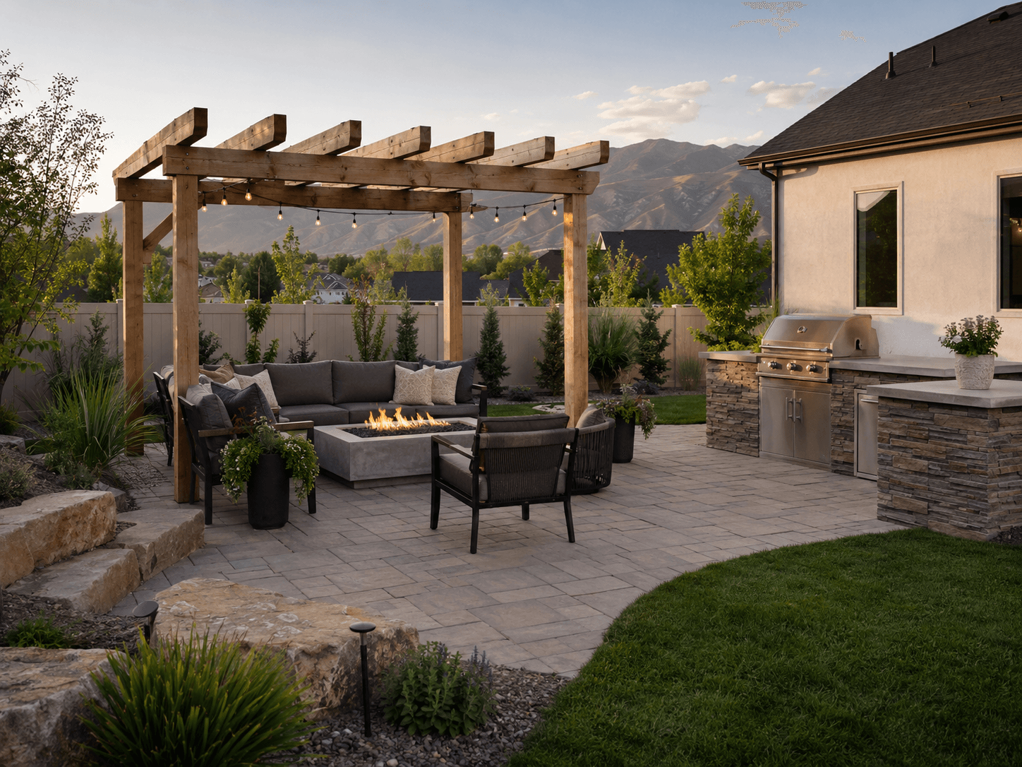 Covered pergola with string lights over a paver patio, gas fire pit, sectional seating, and a stone outdoor kitchen, with the Wasatch foothills behind